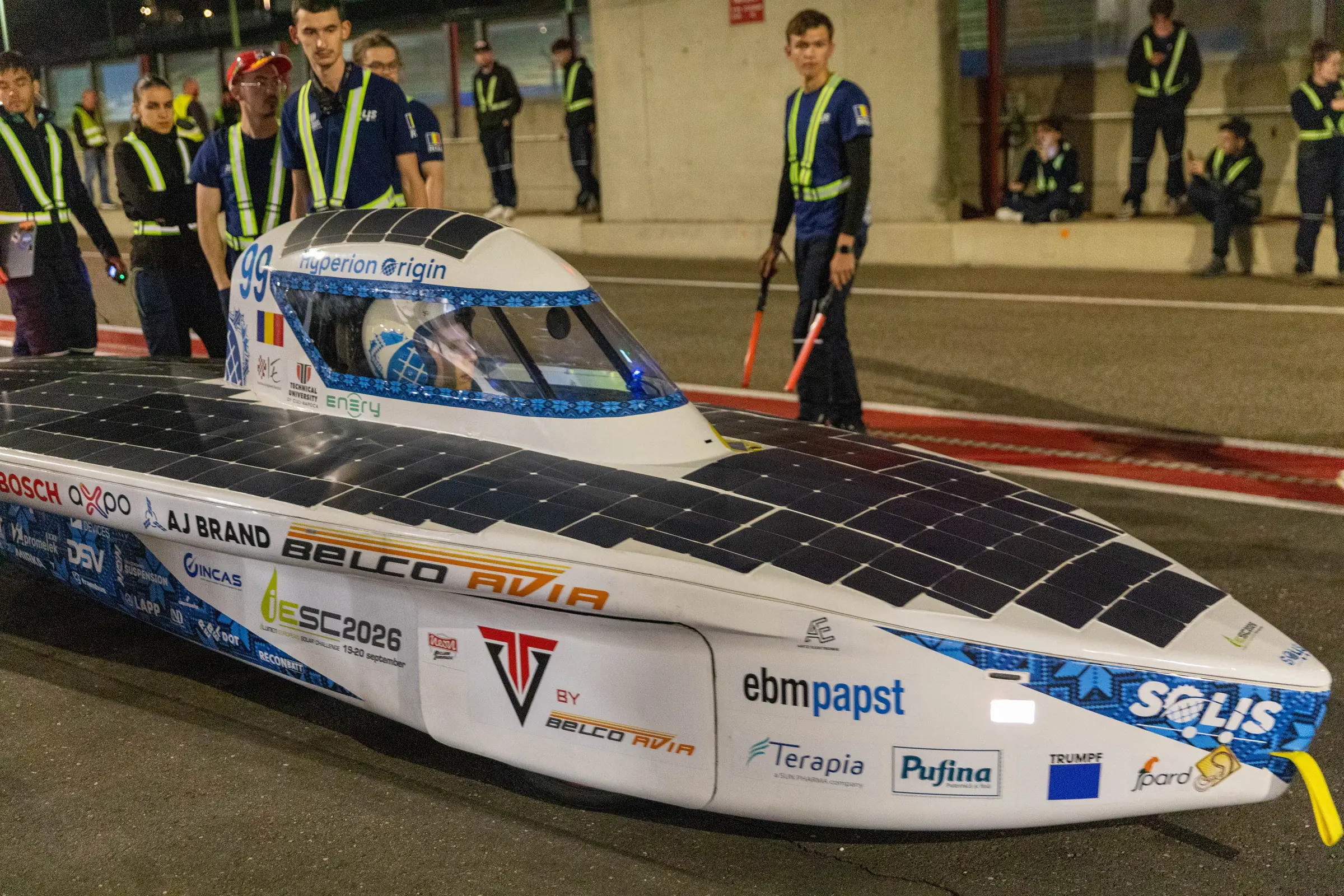 Hyperion Origin under the pit lane lights at night, its side covered in partner logos, with the crew standing around it.