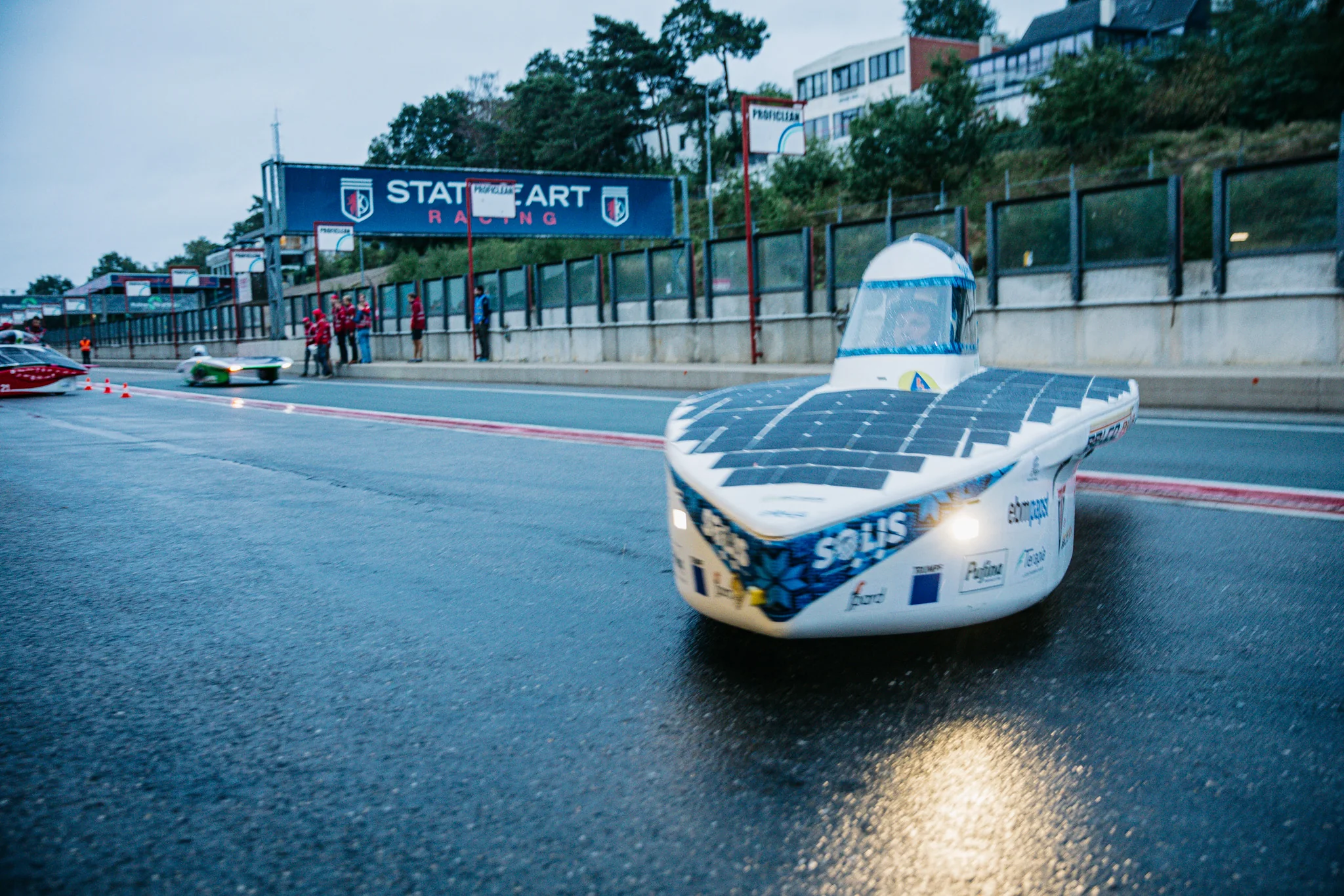 Hyperion Origin coming down a wet track at dawn with its lights reflecting on the asphalt.