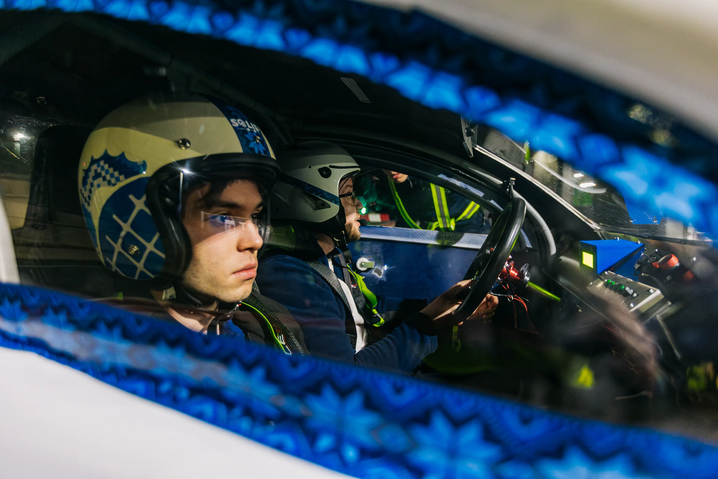 Two helmeted drivers inside the car at night, seen through the window, the blue cross-stitch band around the frame.