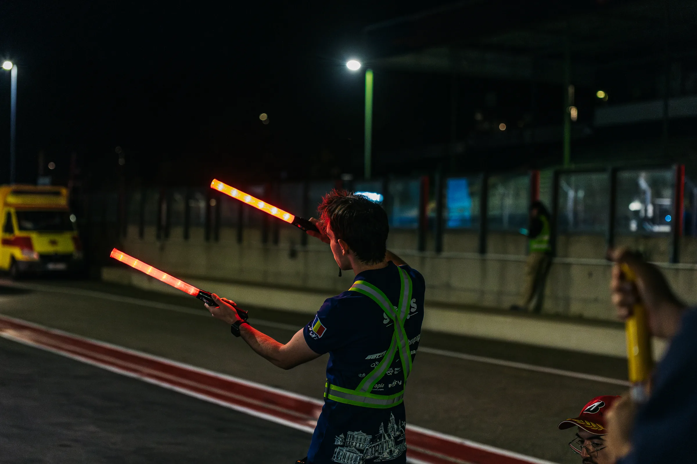 A crew member seen from behind in the dark pit lane, holding two glowing orange light wands.